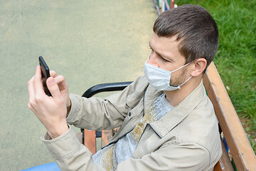 Image showing A man in a protective medical mask sitting on a bench in the park communicates by cell phone