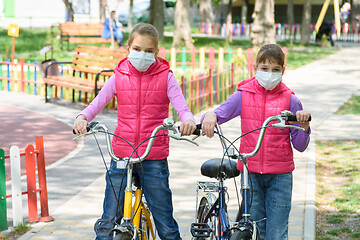 Image showing Two girls with medical masks on their face walk with bicycles in the playground