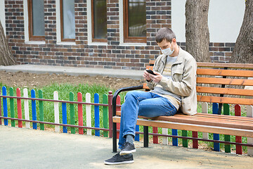 Image showing A man in the isolation mode walks on the street and crouches on a bench, looks at the phone screen