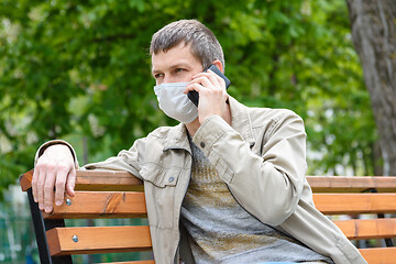 Image showing A man sits on a bench in a medical mask and talks on the phone