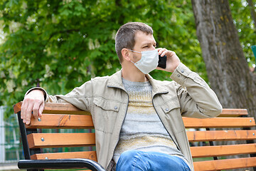 Image showing A man in a protective mask walks through the park and crouches on a bench talking on the phone