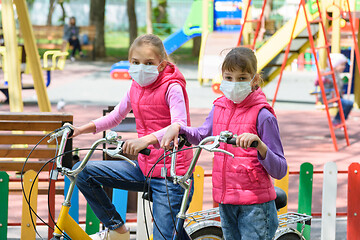 Image showing Two girls in medical masks ride bicycles in the playground