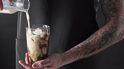 Image showing A male barista with a tattoo pours fresh milk into a glass with ice and coffee.