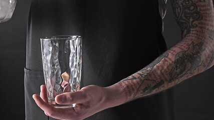 Image showing A man with a tattoo is pouring ice cubes into a glass around a dark background. 4K UHD video, 2160p.