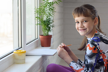 Image showing Happy little girl draws a drawing while sitting on the floor near a large window