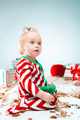 Image showing Cute baby girl 1 year old near santa hat posing over Christmas background. Sitting on floor with Christmas ball. Holiday season.