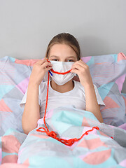 Image showing A girl in a medical mask in quarantine knits and made a smile out of thread on the mask