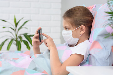 Image showing The girl lies in bed in a medical mask and does web surfing on the phone