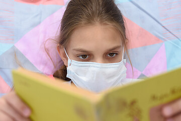 Image showing Girl in a medical mask reads a book while lying in bed, close-up