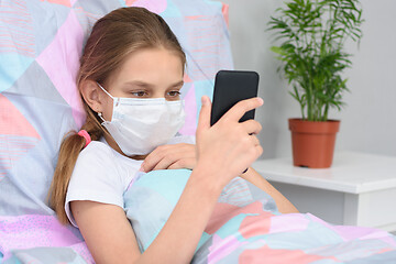 Image showing A girl in a medical mask lies in bed and looks at the phone screen