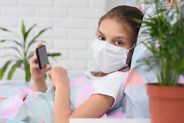 Image showing The girl lies on the bed in a medical mask with a mobile phone and looked into the frame