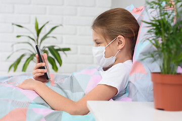 Image showing A girl in a hospital ward lies on a bed in a medical mask and does web surfing on the phone