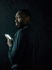 Image showing Studio shot of young serious black African man thinking while talking on mobile phone against black background