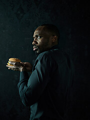 Image showing young african american man eating hamburger and looking away on black studio