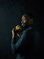 Image showing young african american man eating hamburger and looking away on black studio