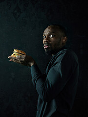 Image showing young african american man eating hamburger and looking away on black studio