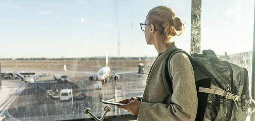 Image showing Young woman standing near airport gates window holding cellphone in her hands, wearing travel backpack and walking to lounge area. Female traveler search online map at the airport