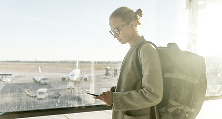 Image showing Young woman standing near airport gates window holding cellphone in her hands, wearing travel backpack and walking to lounge area. Female traveler search online map at the airport