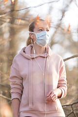 Image showing Portrait of caucasian sporty woman wearing medical protection face mask while walking in park, relaxing and listening to music. Corona virus, or Covid-19, is spreading all over the world