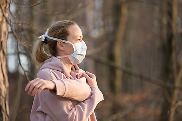 Image showing Portrait of caucasian sporty woman wearing medical protection face mask while relaxing by stretching in forest. Corona virus, or Covid-19, is spreading all over the world