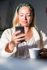 Image showing Beautiful caucasian woman at home, feeling comfortable wearing white bathrobe, taking some time to herself, drinking morning coffee and reading news on mobile phone device in the morning