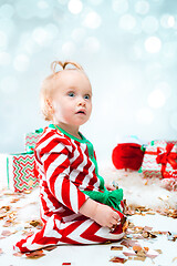 Image showing Cute baby girl 1 year old near santa hat posing over Christmas background. Sitting on floor with Christmas ball. Holiday season.