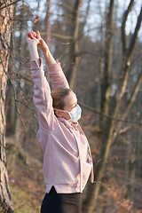 Image showing Portrait of caucasian sporty woman wearing medical protection face mask while relaxing by stretching in forest. Corona virus, or Covid-19, is spreading all over the world