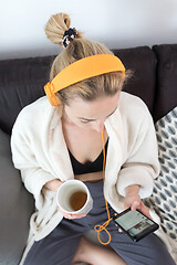 Image showing Stay at home. Social distancing. Woman at home relaxing on sofa couch drinking tea from white cup, listening to relaxing music, stay connected to friens and family via social networks on mobile.