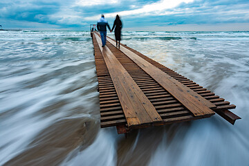 Image showing Two people go down the wooden sea pier into the distance, evening, long exposure