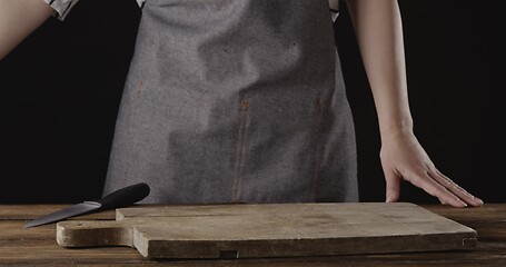 Image showing Chef in a apron are standing at the table with wooden cutting board on it on a black background.