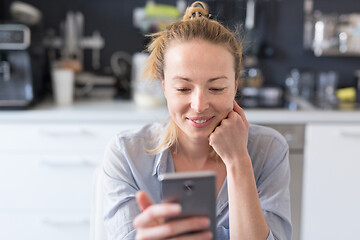 Image showing Young smiling cheerful pleased woman indoors at home kitchen using social media apps on mobile phone for chatting and stying connected with her loved ones. Stay at home, social distancing lifestyle.