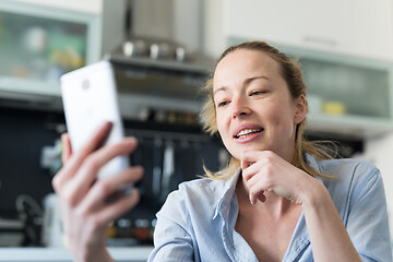 Image showing Young smiling cheerful pleased woman indoors at home kitchen using social media apps on mobile phone for chatting and stying connected with her loved ones. Stay at home, social distancing lifestyle.