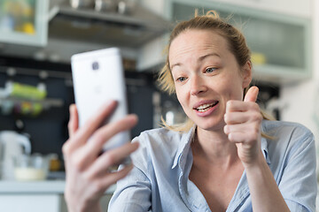 Image showing Young smiling cheerful pleased woman indoors at home kitchen using social media apps on mobile phone for chatting and stying connected with her loved ones. Stay at home, social distancing lifestyle.