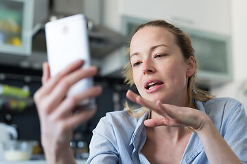 Image showing Young smiling cheerful pleased woman indoors at home kitchen using social media apps on mobile phone for chatting and stying connected with her loved ones. Stay at home, social distancing lifestyle.