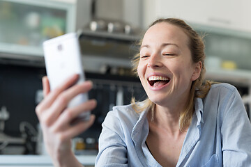 Image showing Young smiling cheerful pleased woman indoors at home kitchen using social media apps on mobile phone for chatting and stying connected with her loved ones. Stay at home, social distancing lifestyle.