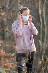 Image showing Portrait of caucasian sporty woman wearing medical protection face mask while walking in park, relaxing and listening to music. Corona virus, or Covid-19, is spreading all over the world