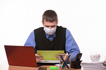 Image showing Office worker in a medical mask at the workplace looks at the tablet screen