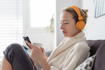 Image showing Stay at home. Social distancing. Woman at home relaxing on sofa couch drinking tea from white cup, listening to relaxing music, stay connected to friens and family via social networks on mobile.