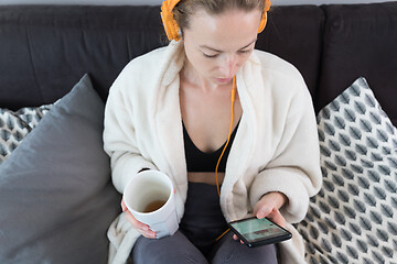 Image showing Stay at home. Social distancing. Woman at home relaxing on sofa couch drinking tea from white cup, listening to relaxing music, stay connected to friens and family via social networks on mobile.