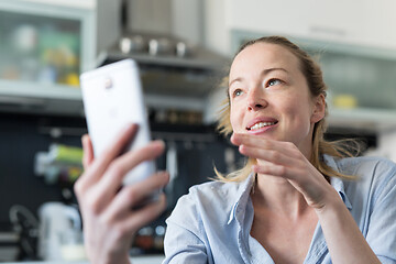 Image showing Young smiling cheerful pleased woman indoors at home kitchen using social media apps on mobile phone for chatting and stying connected with her loved ones. Stay at home, social distancing lifestyle.