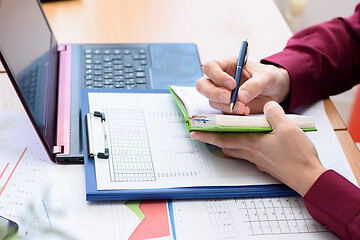 Image showing a person writes in a notebook data from a laptop and documents laid out on the table