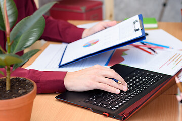 Image showing close-up of hands typing on a laptop on a desk with documents, graphs and tables