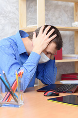 Image showing A man sits at a desk holding his head in a medical mask