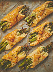 Image showing Baked green asparagus in puff pastry sprinkled with sesame seeds. Placed on a white baking paper