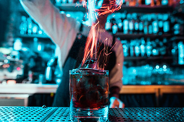 Image showing Glass of fiery cocktail on the bar counter against the background of bartenders hands with fire