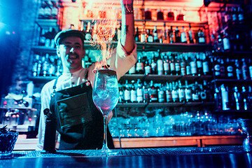 Image showing Glass of fiery cocktail on the bar counter against the background of bartenders hands with fire