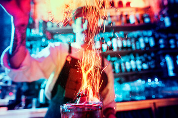 Image showing Glass of fiery cocktail on the bar counter against the background of bartenders hands with fire