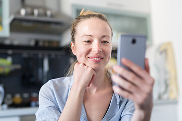 Image showing Young smiling cheerful pleased woman indoors at home kitchen using social media apps on mobile phone for chatting and stying connected with her loved ones. Stay at home, social distancing lifestyle.