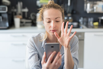 Image showing Young pleased woman indoors at home kitchen using social media apps on mobile phone for chatting and stying connected with her loved ones. Stay at home, social distancing lifestyle.