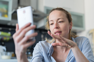 Image showing Young pleased woman indoors at home kitchen using social media apps on mobile phone for chatting and stying connected with her loved ones. Stay at home, social distancing lifestyle.